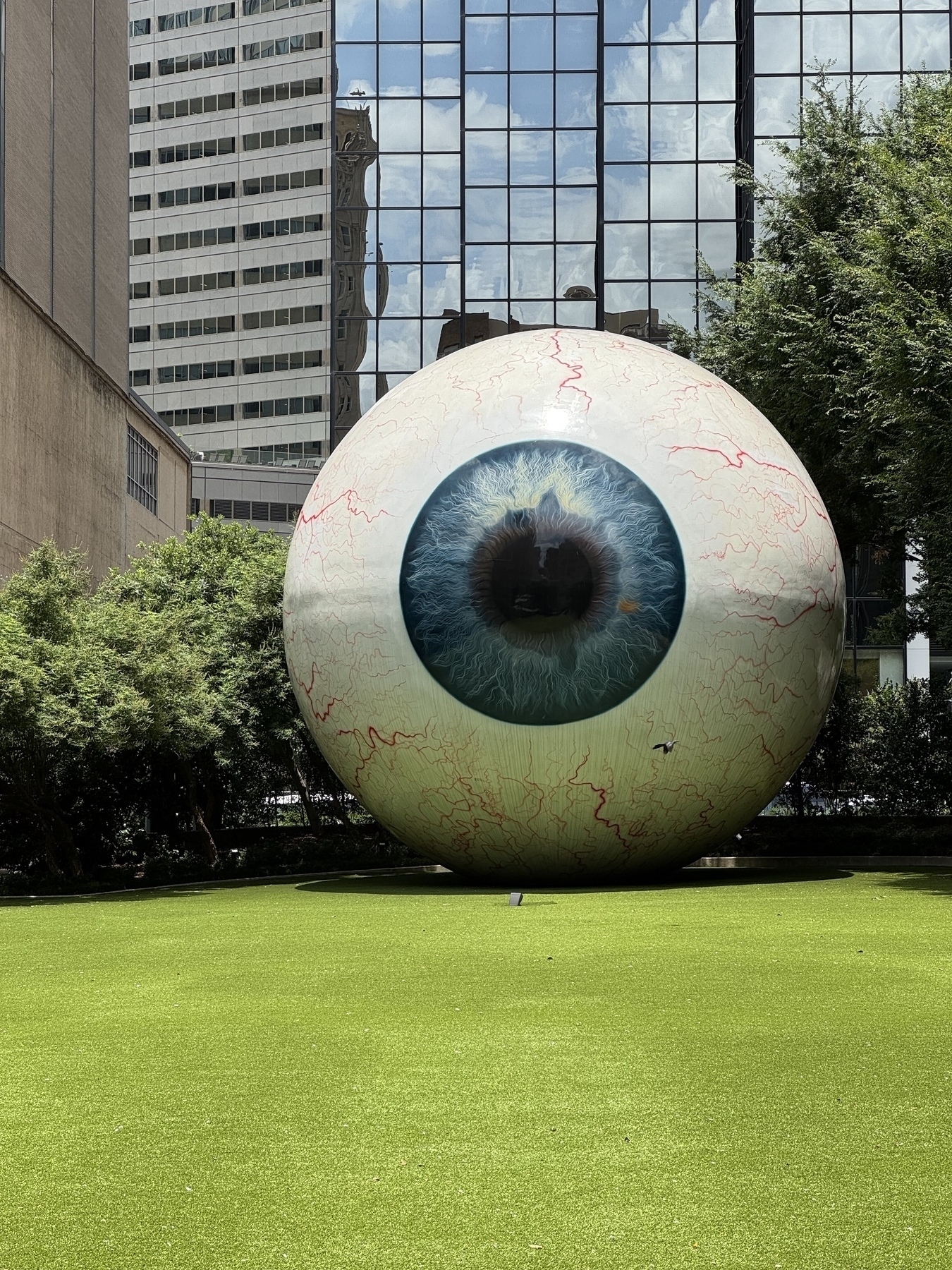 A large, realistic sculpture of an eyeball is displayed on a patch of grass surrounded by urban buildings and trees.