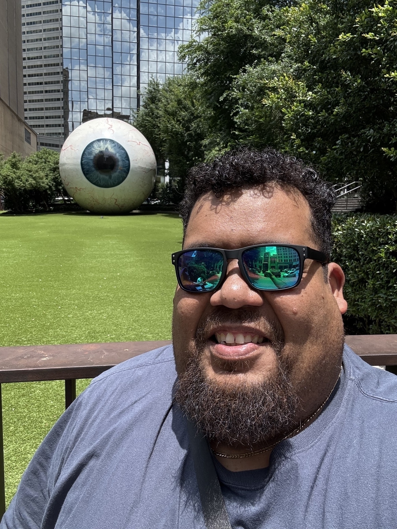 Me with sunglasses smiles for a selfie in front of a large, decorative eyeball sculpture on a grassy area.