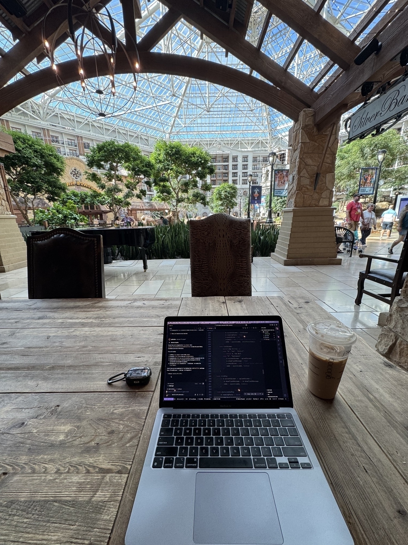 A laptop and iced coffee sit on a wooden table in a spacious indoor area with a high glass ceiling and lush greenery.