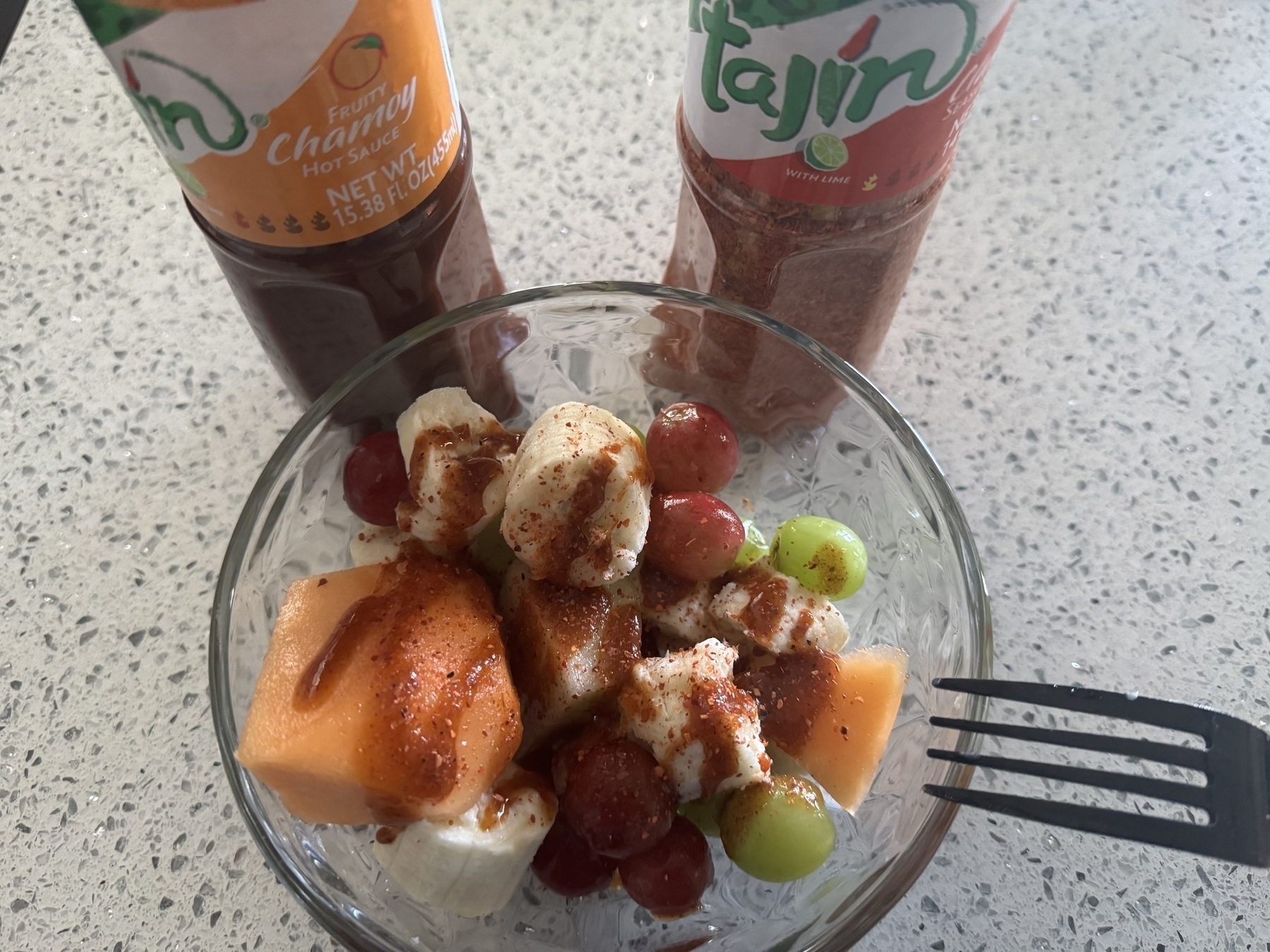 A bowl of fruit topped with Chamoy and Tajín seasoning is accompanied by bottles of both condiments on a speckled countertop.