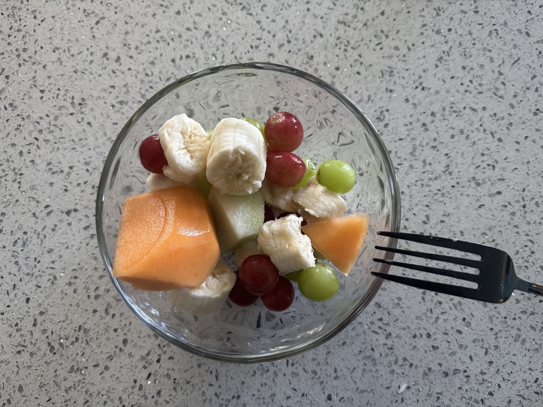 A glass bowl contains a fruit salad with bananas, grapes, cantaloupe, and honeydew melon on a speckled countertop, accompanied by a fork.